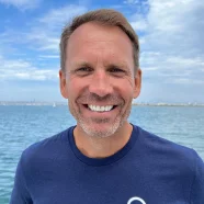 Smiling man in a blue shirt stands by the water with a cloudy blue sky in the background.