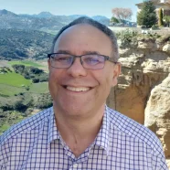 Smiling person in glasses and checked shirt standing in front of a scenic cliffside and valley landscape.