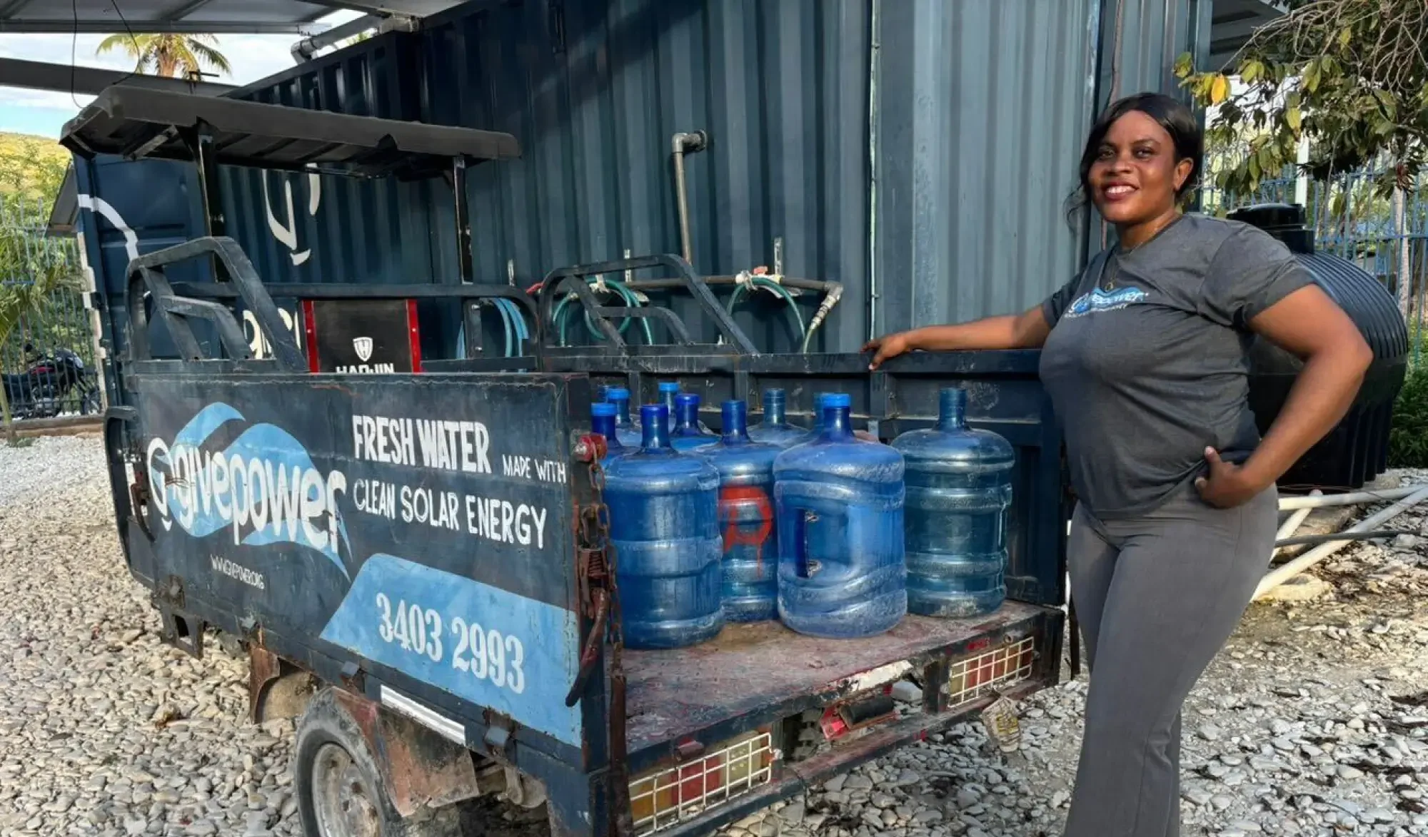 Una mujer está de pie junto a una camioneta pequeña en la que hay jarras de agua y un letrero que dice: “Agua potable con energía solar limpia”.