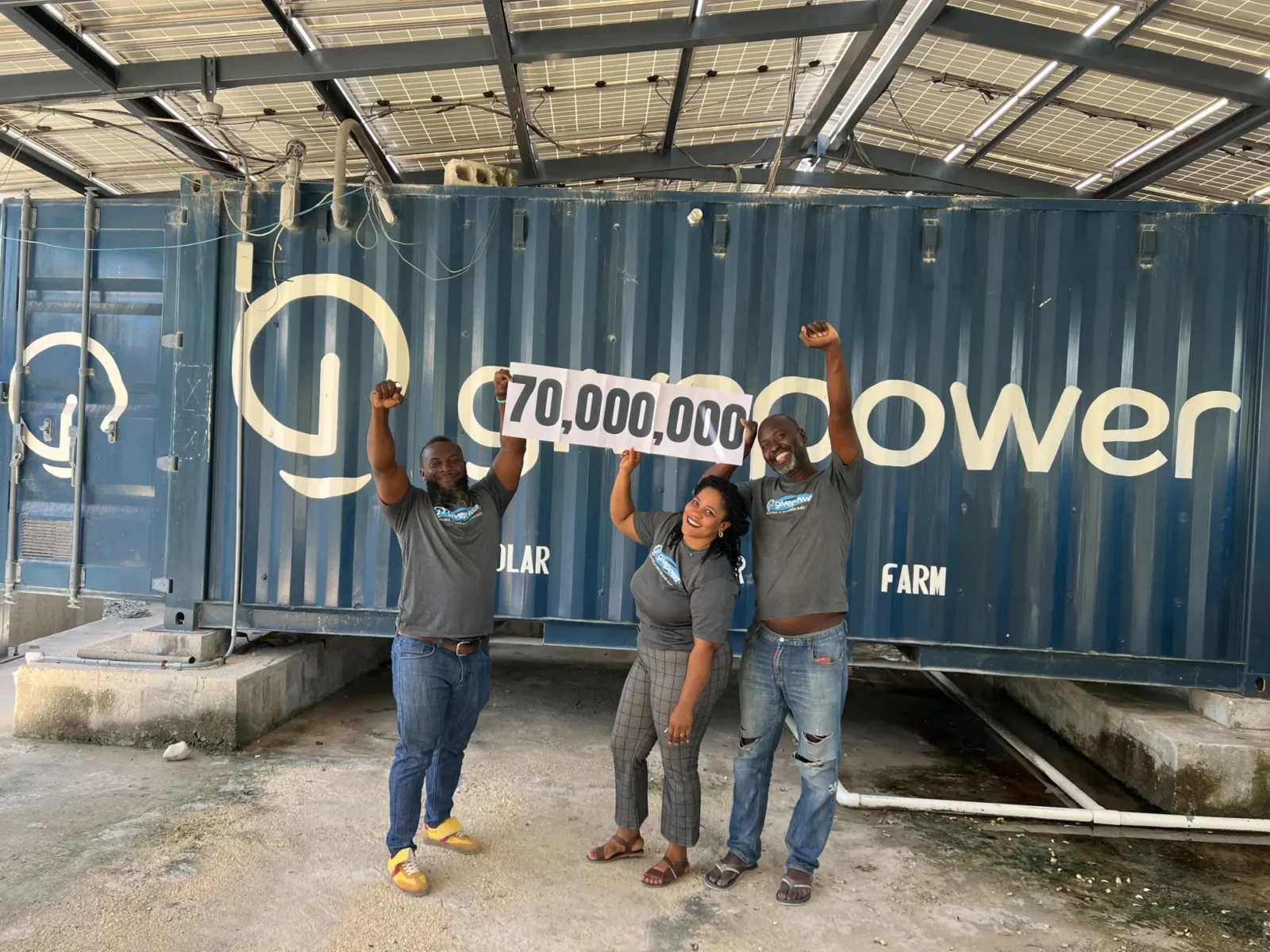 Tres personas celebran sosteniendo un letrero con el número 70 000 000 frente a un contenedor azul con la inscripción «O...power...».