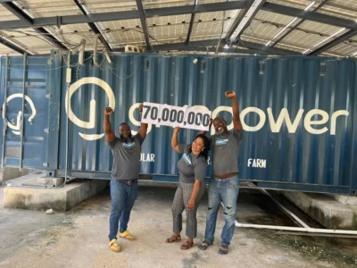 Tres personas celebran sosteniendo un letrero con el número 70 000 000 frente a un contenedor azul con la inscripción «O...power...».