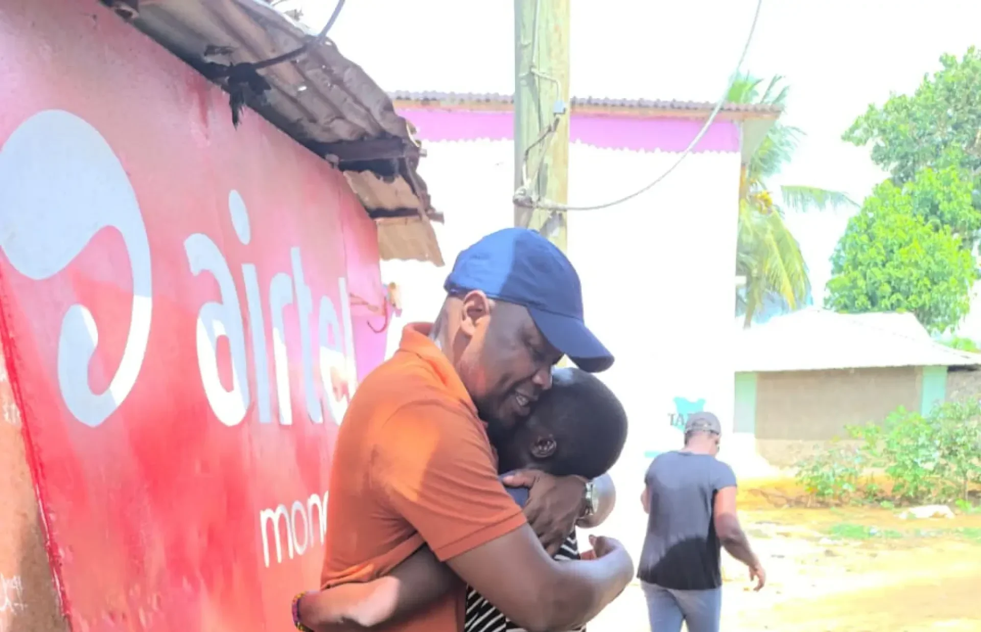 A man and a woman share a warm hug on a sandy street near a red wall, while others walk by.