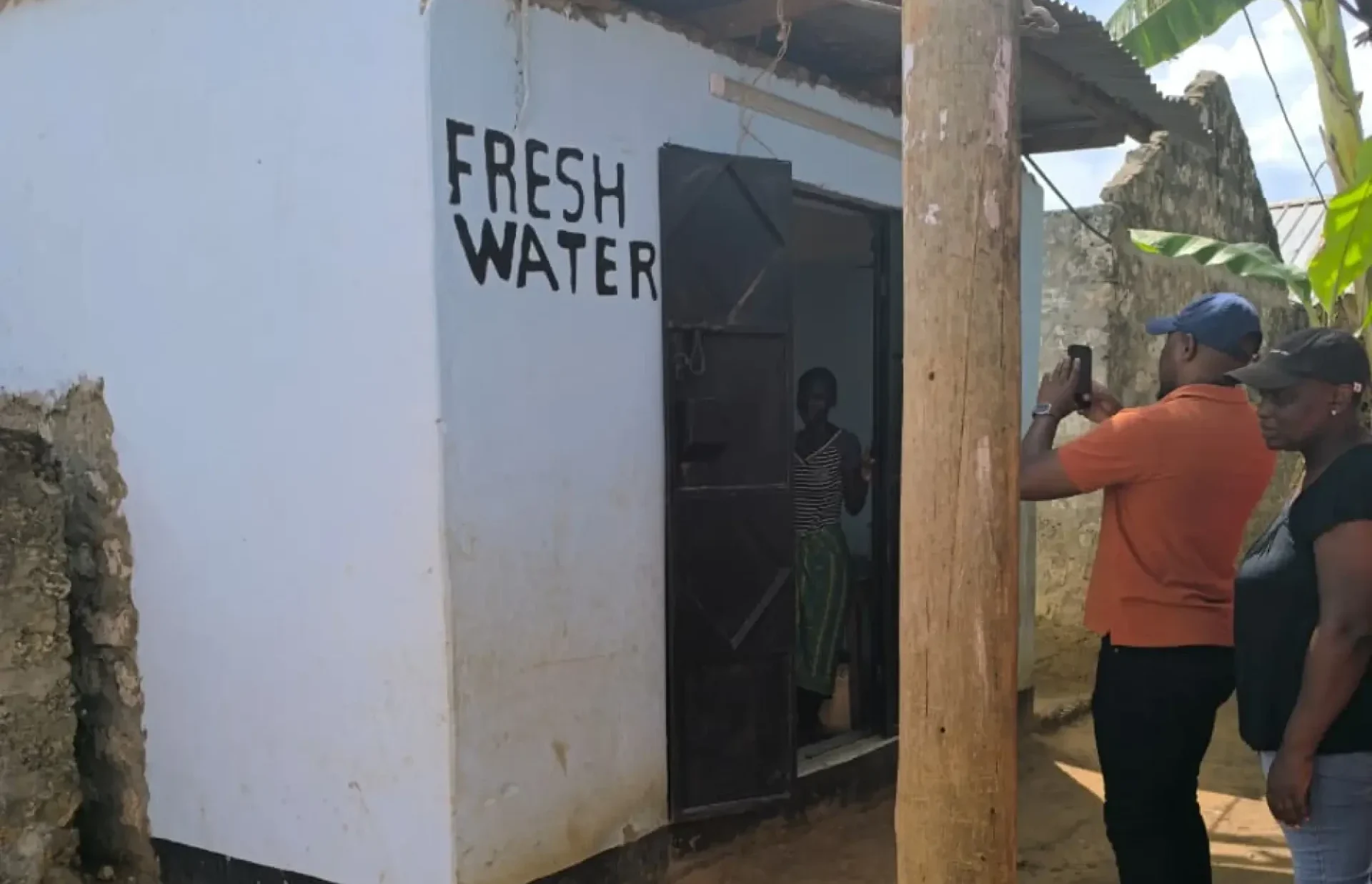 Three people stand by a small building labeled FRESH WATER, with one person taking a photo at the door.