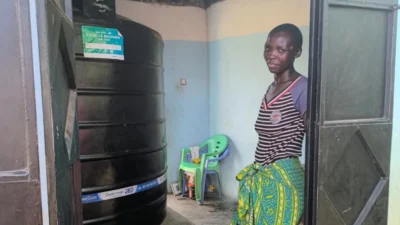 Una mujer de pie en el umbral de una puerta, con un gran tanque de agua negro y sillas de plástico verdes dentro de la habitación.