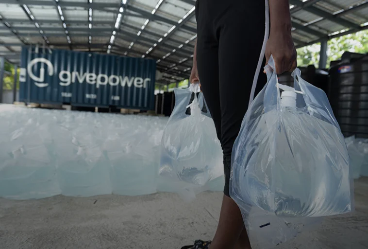 Person holding large bags of water in a facility with a givepower container and many water bags.