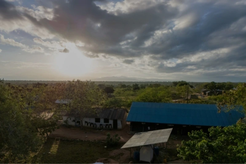Sunset over solar system, rural buildings with blue rooftops, surrounded by trees under a cloudy sky.