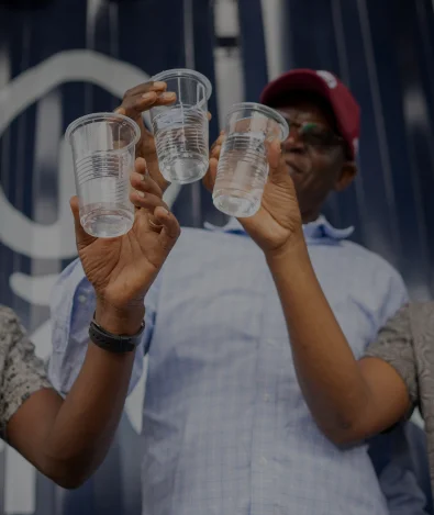 Two people holding up clear plastic cups of water in front of a dark blue background, faces partially visible.