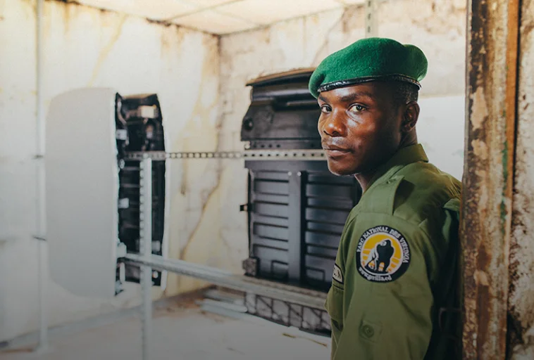 A man in a green uniform and beret stands indoors, with equipment and a wall in the background.