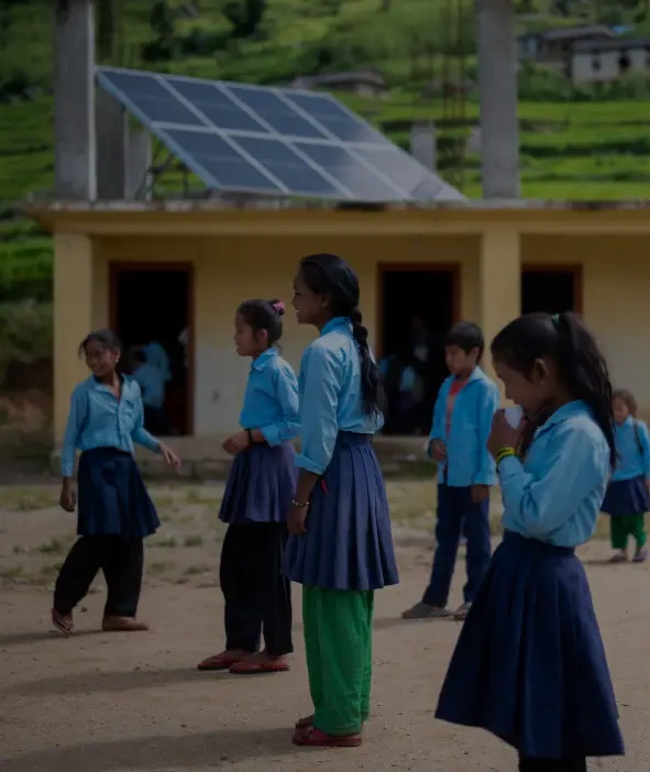 Unos niños con uniformes escolares están parados frente a un edificio con paneles solares en el techo, en un entorno rural.
