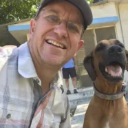 Man wearing glasses and a cap smiles next to a happy brown dog outdoors on a sunny day.