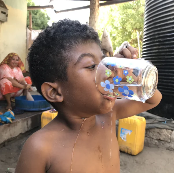 A young boy drinks water from a glass, with water spilling down his chin. People and containers are in the background.
