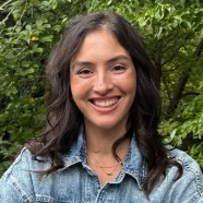 Woman with long dark hair smiles outdoors, wearing a denim jacket with green foliage in the background.