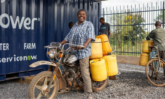 A smiling man on a motorbike with yellow water containers; two people and a blue container are in the background.