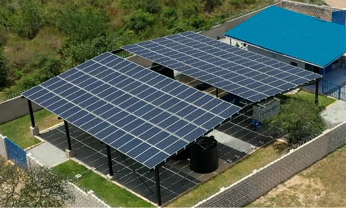 Large solar panels installed on metal frames beside a blue-roofed building, surrounded by trees and walls.
