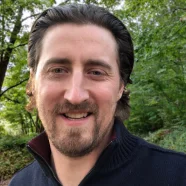 Man with brown hair and a beard smiles outdoors with green trees in the background.