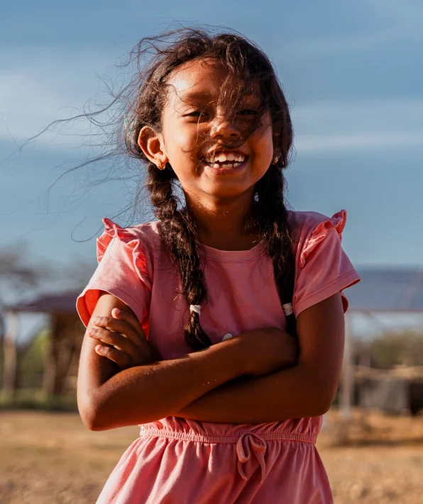 Una niña sonriente, con el cabello trenzado y un vestido rosa, está de pie al aire libre, en un lugar donde la energía solar prospera para las comunidades remotas.