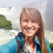 Smiling woman in a blue jacket stands in front of a large waterfall with mist and greenery.