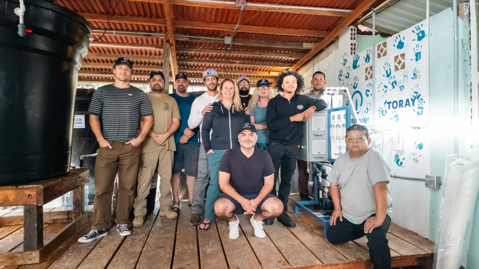 Eleven people pose together inside a rustic building with tanks, pipes, and a Toray water system.