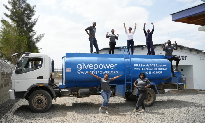 Seven people pose on and around a blue water truck with givepower and fresh water made with clean solar energy text.