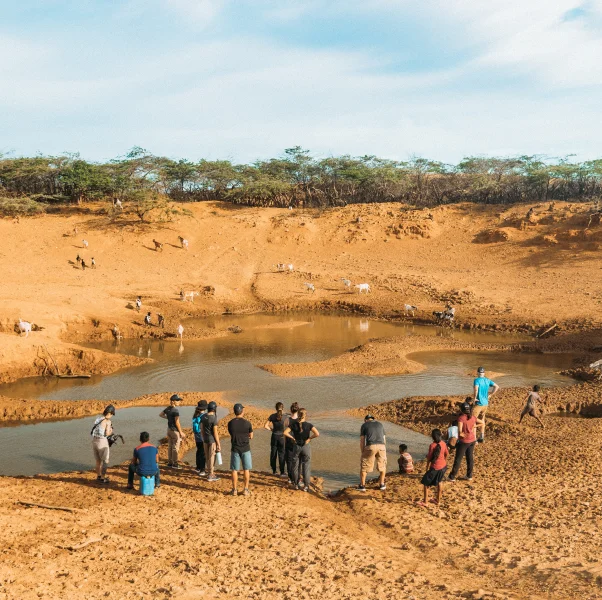 People gather around a small muddy pond in a dry landscape with sparse trees in the background.