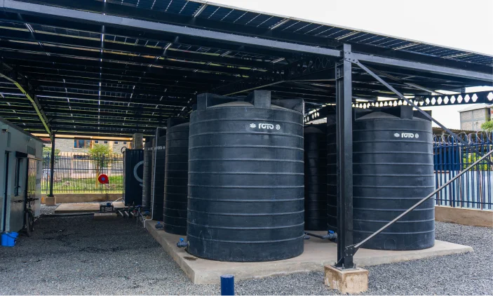 Four large black water storage tanks sit under a metal canopy in an outdoor, fenced area.
