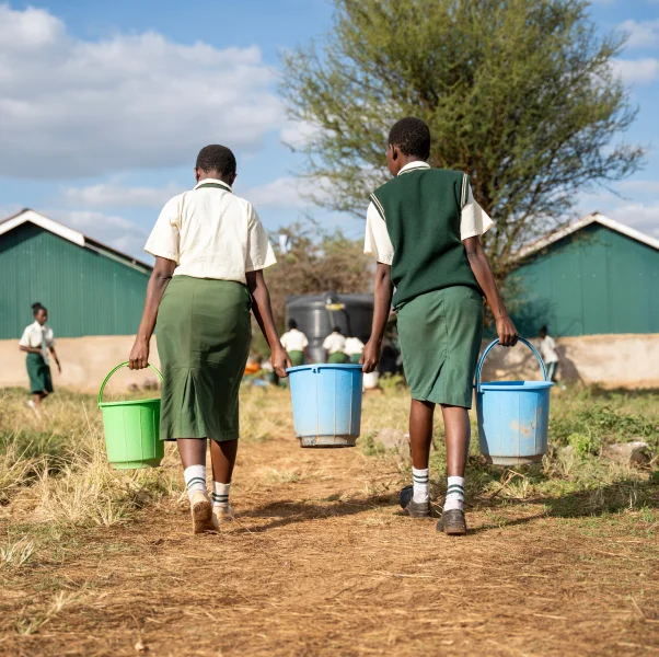 Two students in uniforms carry plastic buckets along a dirt path at a schoolyard on a sunny day.