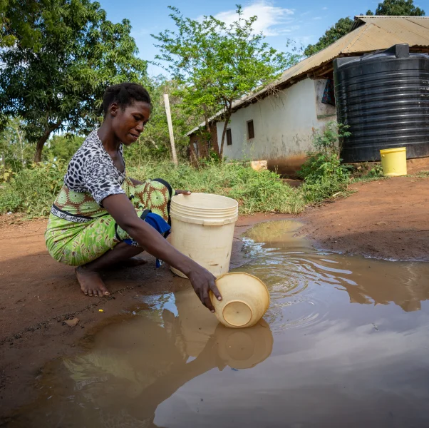 Woman filling a bucket with water from a muddy puddle outside a house in a rural area.